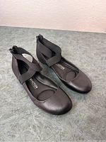 Pair of black Kenneth Cole Reaction ballet flats with criss-cross elastic straps and back zipper closure, shown on grey surface from angled top view.