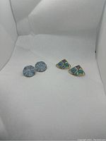 Two pairs of pastel-colored clip-on earrings on white background: triangular pair with enamel insets and blue round pair.