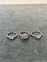 Three silver-tone rings displayed on a textured gray surface, showing different stone styles and sizes.
