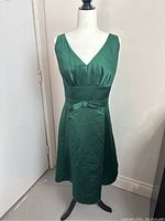 Full front view of sleeveless green satin dress on a mannequin, showing V neckline, fitted waistband with bow, and knee-length A-line skirt.