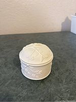 Front angle view of the small round bisque trinket box showing embossed cherry blossom floral pattern on matte white surface.