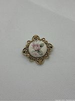 Front view of vintage gold-tone metal brooch with central porcelain floral design showing pink rose and green leaves on white background.