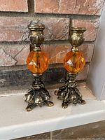 Front view of two vintage Hollywood Regency orange faceted candlestick holders with brass ornate bases and tops in front of a brick wall.