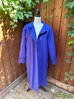 Full length view of the vintage oversized trench coat displayed on a dress form outdoors, showing royal blue/lilac color, wide collar, pockets, and button closure.