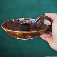 Close-up side view of glazed brown vintage candle holder with circular base, loop handle, and central candle socket held in hand against green background.