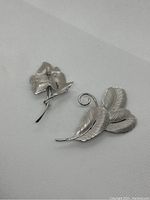 Two sterling silver brooches, a floral design and a leaf design, laying on white textured fabric.