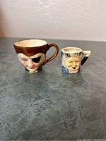 Photo showing two vintage Toby mugs on a grey surface: one larger pirate-themed mug and one smaller elderly man face mug.