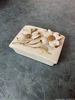 Closed view of vintage white ceramic trinket box with raised white flowers and gold accents on lid.
