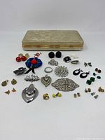 Full view of costume jewelry laid out in front of vintage textured jewelry case. Shows multiple earrings, brooches, pendant, scarf clip, and pin assortment.