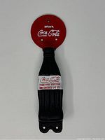 Full view of the black cast iron door handle shaped like a Coca-Cola bottle with a painted white and red label and red circular mounting plate with Coca-Cola logo.