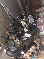 Photo showing multiple candlesticks of assorted materials including glass, brass, and pewter, arranged on a wooden table.