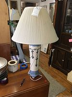 Full view of ceramic table lamp on wood surface showing blue floral vine pattern on white base, and cream pleated pleated lampshade.