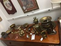 Photo showing various copper and brass items on a wooden table including bowls, pitchers, small containers, and a brass Buddha figure.