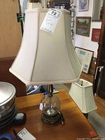 Full view of the lamp showing the crackle glass ball base, dark metal base, power cord, and large beige fabric shade.