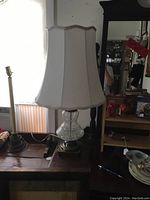 Table lamp with crackle glass base and white fabric shade placed on a wooden table beside other furniture.
