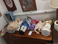 Seven dolls displayed on a wooden surface including a boxed Shirley Temple collectible and six loose dolls in various outfits.