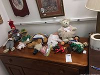 Wide view of the assortment of dolls including teddy bear in pink tutu, male doll in red hat, traditional dressed female doll, and other small dolls on a wooden table.