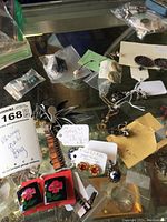 Photo shows assorted earrings laid on a reflective surface with several pairs visible including floral black and red, silver leaf clip-ons, dangle earrings with copper links, small stud earrings with pearls and rhinestones.
