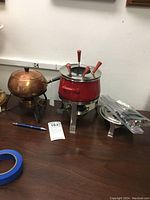 Photo showing two fondue pots; one copper and one red with a set of colorful forks wrapped in plastic beside them.
