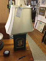 Full lamp showing green wooden base with floral decoration and white pleated fabric shade.