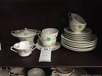 Photo showing a partial set of Haviland china pieces on a wooden shelf including teacups stacked on saucers, a covered serving dish, creamer, and a stack of small plates.