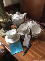 Large coffee pot, sugar bowl, cups, saucers, and plates arranged on table with a pen and handwritten pricing note showing item details and price.