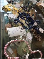 Photo showing mixed jewelry pieces on a reflective surface, including blue lapis chip necklace with earrings, gold-tone chain bracelet, and pink and white beaded necklace.