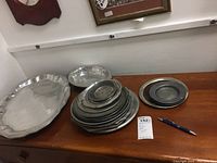 Wide view of the pewter serving platter and stacked plates on a wooden surface with a pen for scale.