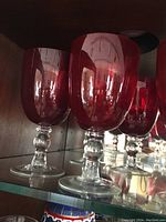 Close-up image of five large water goblet glasses with vibrant red bowls and short clear stems arranged on a shelf.