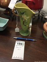 Front and overall view of the green Roseville pottery vase with apple blossom relief and bamboo-like handle alongside a pen and maxsold lot tag