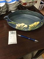 Blue oval Roseville pottery bowl with yellow and white freesia flower decorations on the sides with handles. Pen and lot number card included for scale.