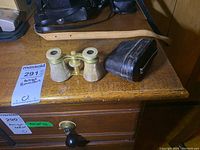 Antique small opera glasses with mother of pearl trim and brass, alongside black leather case on wooden table with leather strap visible.