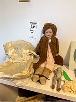 Full view of the antique porcelain head doll seated, wearing brown coat and hat, pink dress, small porcelain doll beside it, and fabric pieces on a white surface