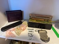Photo of assorted vintage sewing and personal items on white surface with green tape edge, including antique sewing box, Monarch rug needles, handkerchief box, miniature purse, eyeglasses, and hair net.