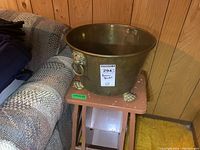 Brass bucket placed on a stool in front of wood panel wall, showing front and one side with lion head handle and claw feet.