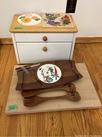 Wood cutting board, wooden serving plate with ceramic insert, carved salad servers, and cheese knife and fork arranged on a wooden surface.