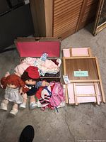 Photo showing two Cabbage Patch dolls, a pink and wood doll crib, and various doll clothes including some in a box
