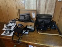 Photo showing assortment of vintage cameras, camera cases, and stereo headphones arranged on wooden dresser with wood panel wall background.