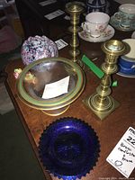 Photo showing a pair of brass candlestick holders, a large glass bowl with a dated note inside, and additional glass bowls and porcelain teacups in the background.