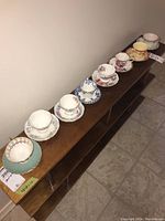 All 8 assorted china cups and saucers lined up on wooden shelf, showing variety of patterns and colors including floral designs and gold trim.