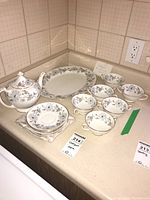 Full view of the tea set laid out on a kitchen countertop showing 6 cups and 6 saucers, teapot with lid, and the platter along with MaxSold tags and seller's handwritten sign confirming Coalport China My Fair Lady pattern.