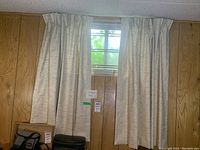 First photo showing one set of beige curtain panels installed on a basement window with wood-paneled walls.