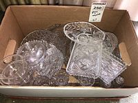 Wide shot of the entire lot showing a cardboard box filled with various glass and crystal serving dishes including a covered dish and multiple plates and trays.