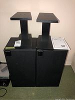 Pair of KEF K Series speakers shown with their black stands placed on the floor, against a wall, with a manual on top of one speaker. Speakers have fabric covers and stand plates are rectangular.
