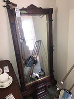 Full view of vintage wood framed mirror standing against wall, showing carved upper decorative molding and metal brackets on frame corners.