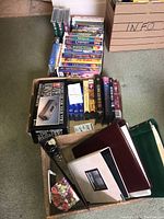 Overall view of many VHS tapes stacked in cardboard boxes featuring colorful Disney titles and other movies, also showing VHS binders and a VHS rewinder device in packaging.