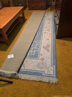 Photo showing the full length of the rolled-out rug along with beige fringe at the visible end, placed beside wooden furniture on a mustard carpet.