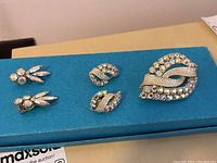 Four pieces of jewelry displayed on blue background: two pairs of earrings and one brooch, showing aurora borealis rhinestones and gold-tone metal