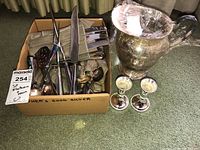 Box containing various silver plated flatware and serving utensils along with two candlestick holders and large silver pitcher.