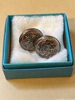 Pair of sterling silver screwback earrings resting on white padding inside a blue box, showing the round shape and engraved floral design.